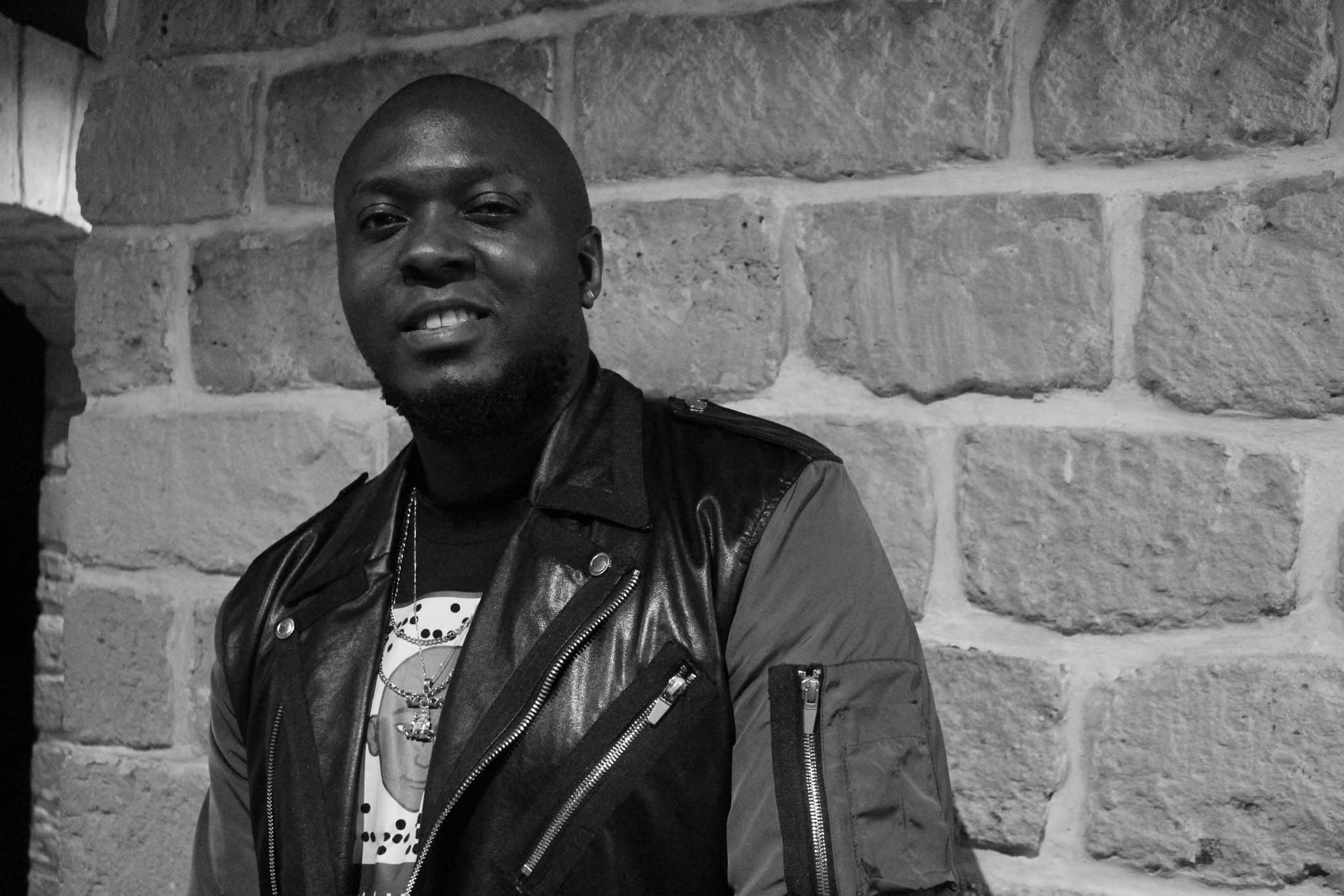 Black Moz en portrait devant un mur en pierre, veste en cuir, noir et blanc
